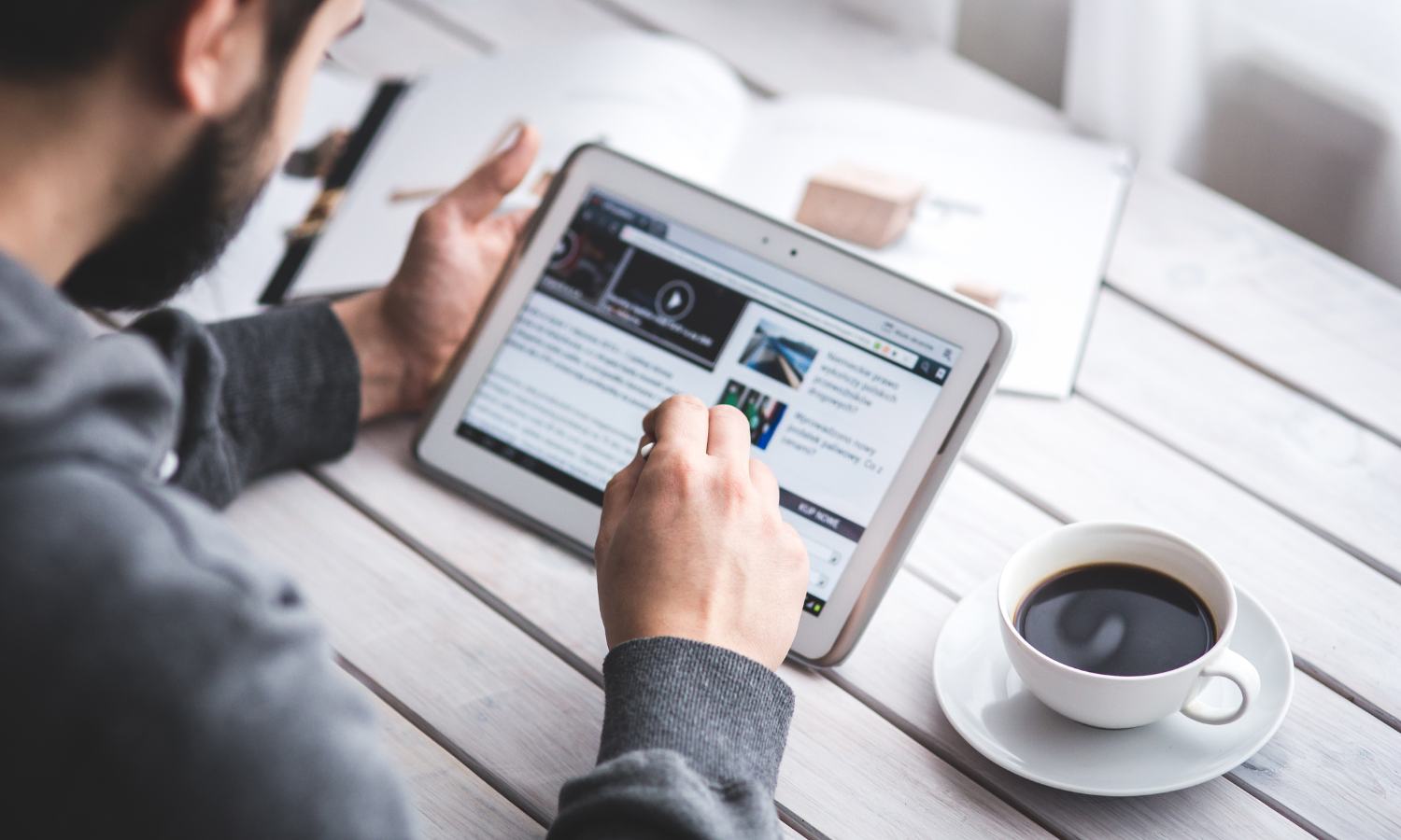 The photo angle provides a look over a man's shoulder to the table where he is seated. He is reading an online article on a tablet. To his left is an open book. To his right is a cup of black coffee.