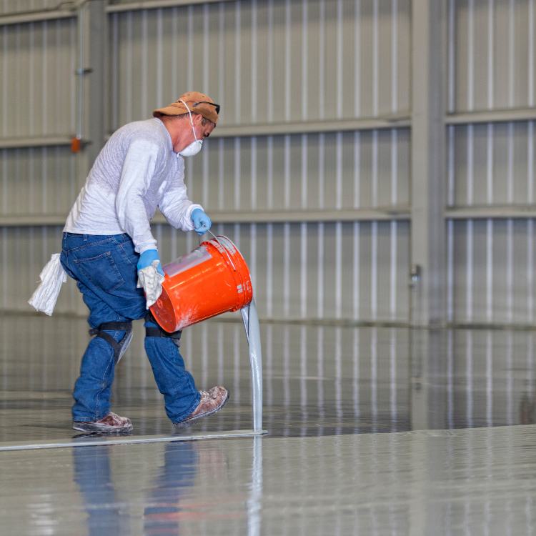 A man in work clothes wears safety gear (gloves, mask, knee pads) as he pours a clear coating from an orange industrial bucket over what appears to be a concrete floor.